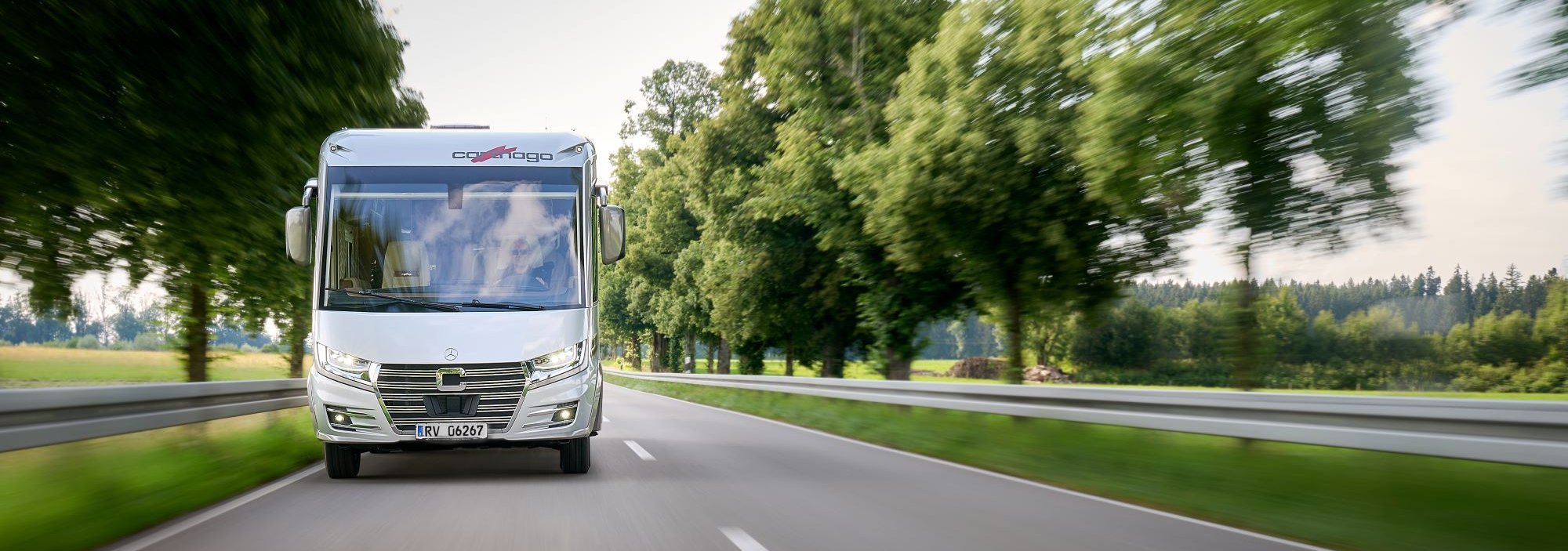 Ansicht eines Carthago Wohnmobils auf einer Landstraße zwischen Bäumen und Wiesen.